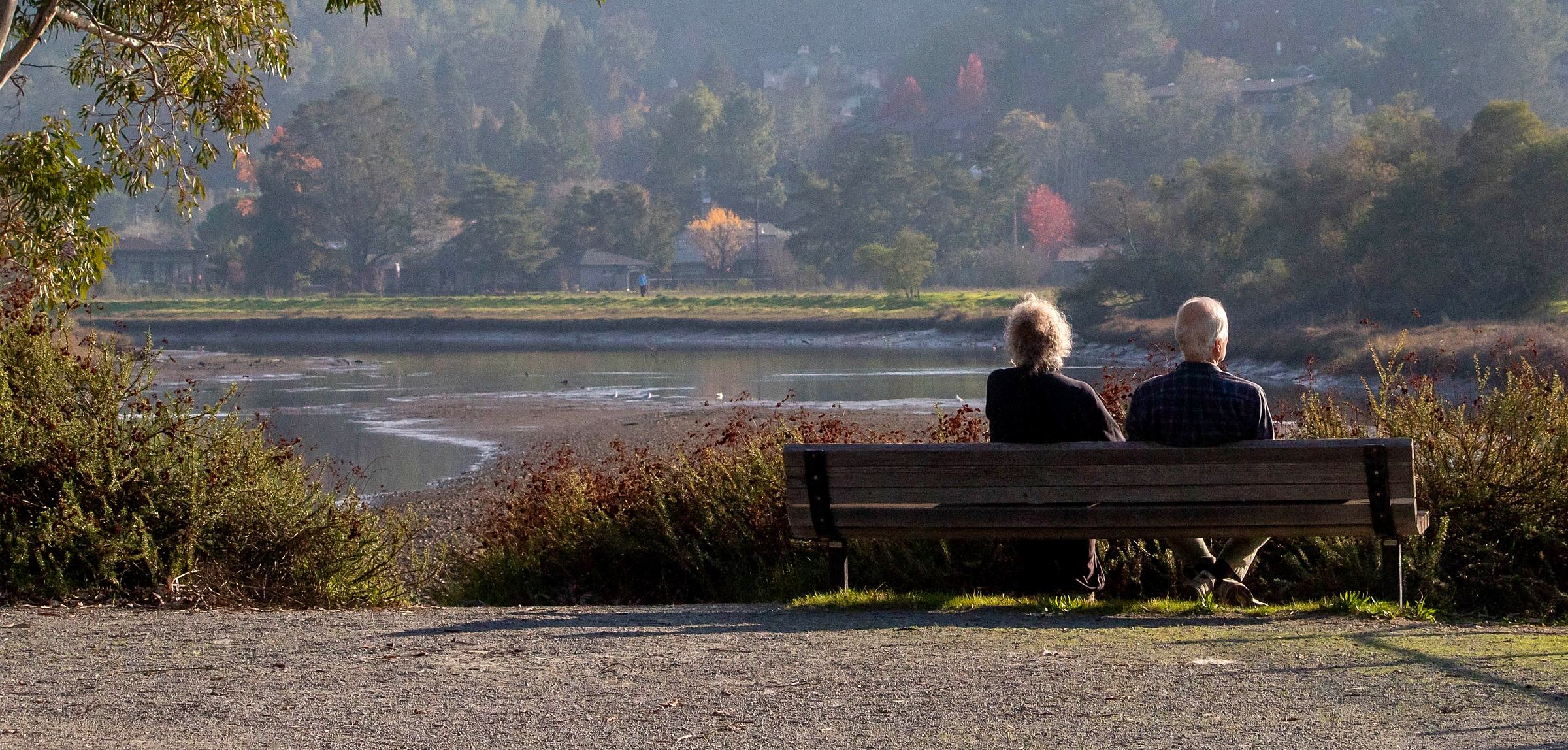 A retired couple sitting on a bench looking out over a quiet inlet